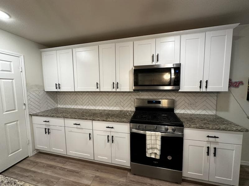 Kitchen with white cabinets, granite countertops, stainless steel stove and microwave, and herringbone tile backsplash.