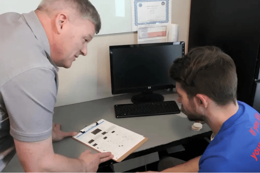 Two men review documents on a clipboard at a desk with a computer monitor and keyboard.