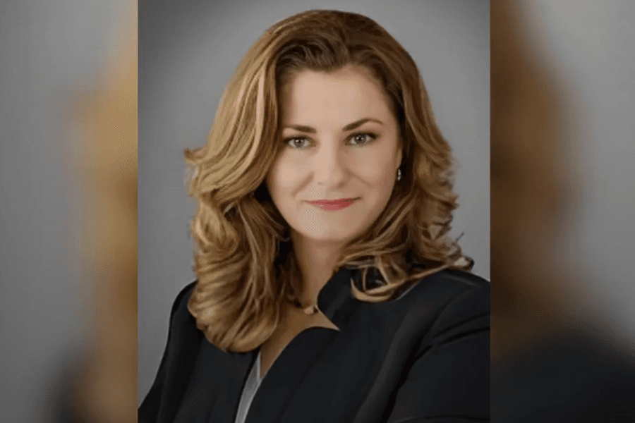 Professional headshot of a woman with wavy blonde hair wearing a black blazer against a gray background.