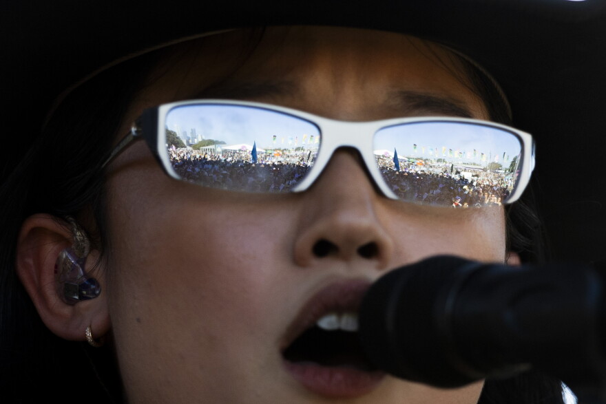 A crowd is reflected in the sunglasses of a woman performing on a stage as she sings into a microphone.