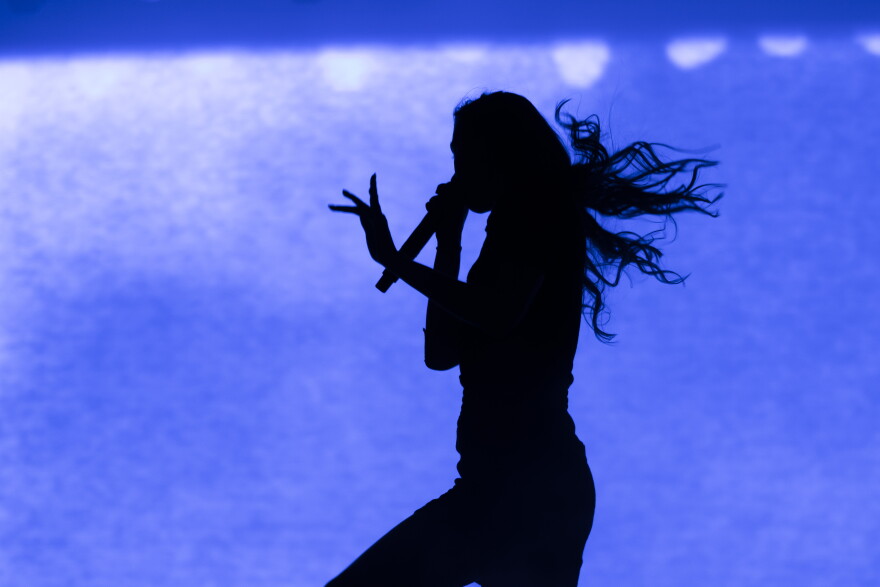 The silhouette of a woman is performing on a stage is seen against a blue backdrop.