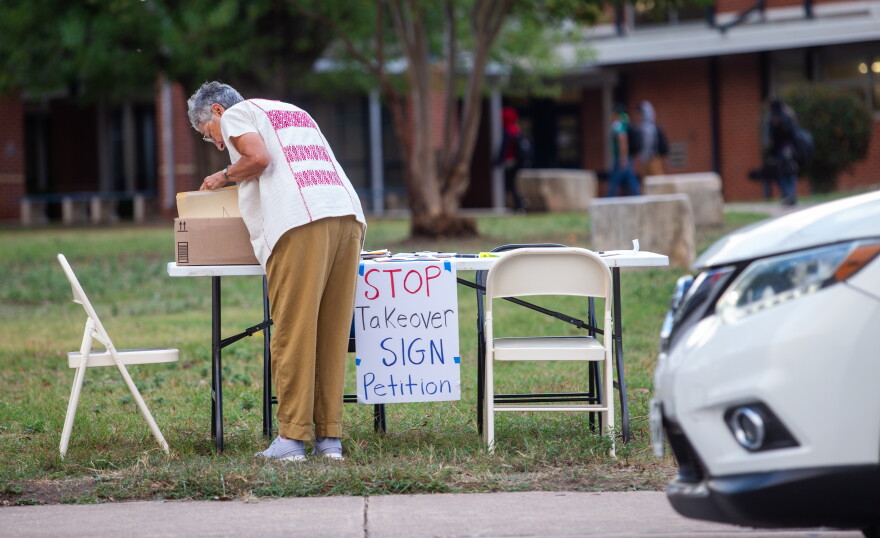 A woman outside of a school, looking into a box with a sign next to her that reads "Stop takeover sign petition."