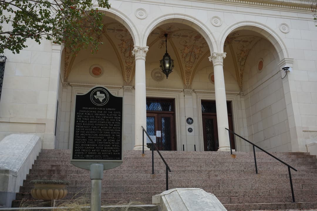 Arched stone entrance of the 1933 Austin Public Library building with a Texas historical marker in front of granite steps.