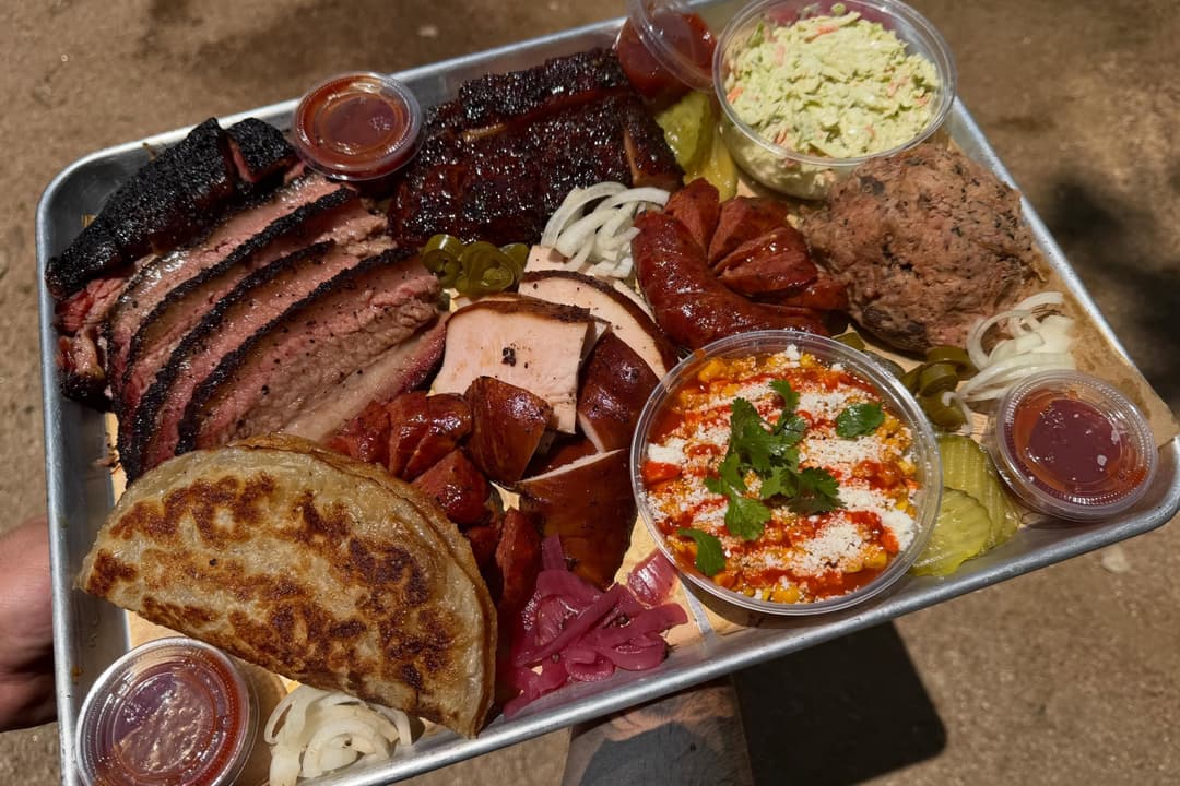 Barbecue platter with sliced brisket, ribs, sausage, turkey, coleslaw, elote, pickles, onions and sauces.