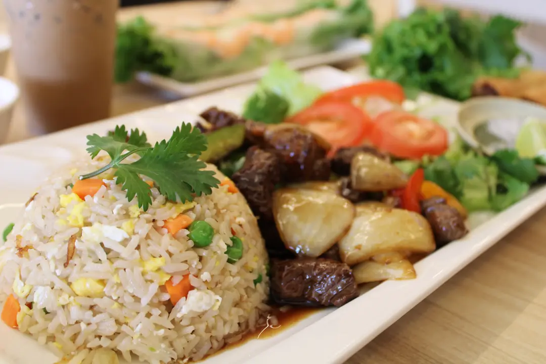 Fried rice with egg and peas served with cubed beef, onions, tomato slices and lettuce on a white plate.