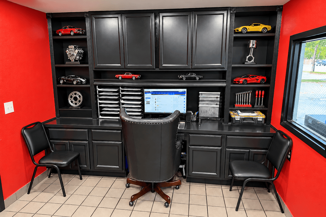 Automotive office with black cabinetry, red walls, desk with monitor, leather chair, and car models on shelves.