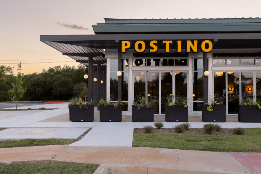 Exterior of Postino restaurant at dusk with glass walls, black planters, and yellow signage.