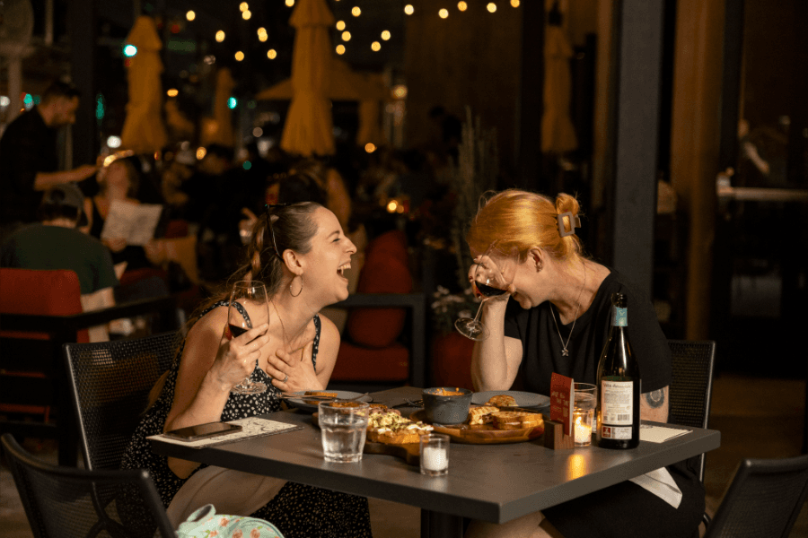 Two women laughing while dining with wine at an outdoor restaurant lit by string lights at night.