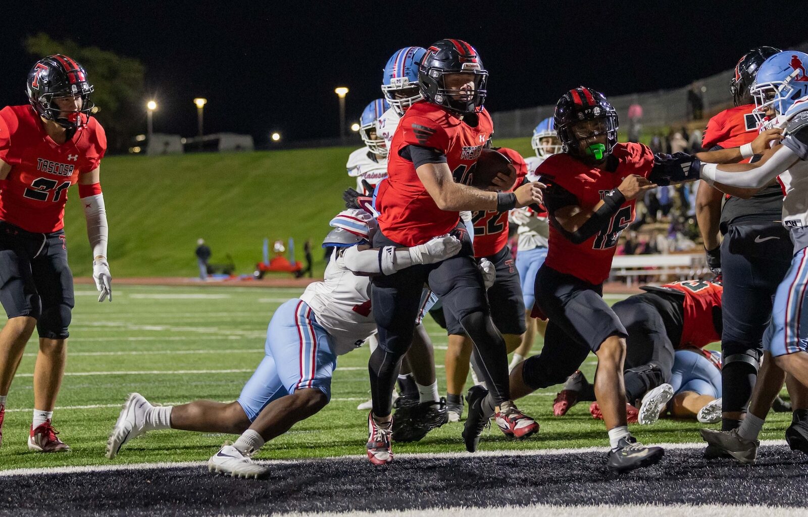 Running back in a red jersey salvages the ball and charges forward as defenders close in during a night football game on a lit field.