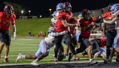 Running back in a red jersey salvages the ball and charges forward as defenders close in during a night football game on a lit field.