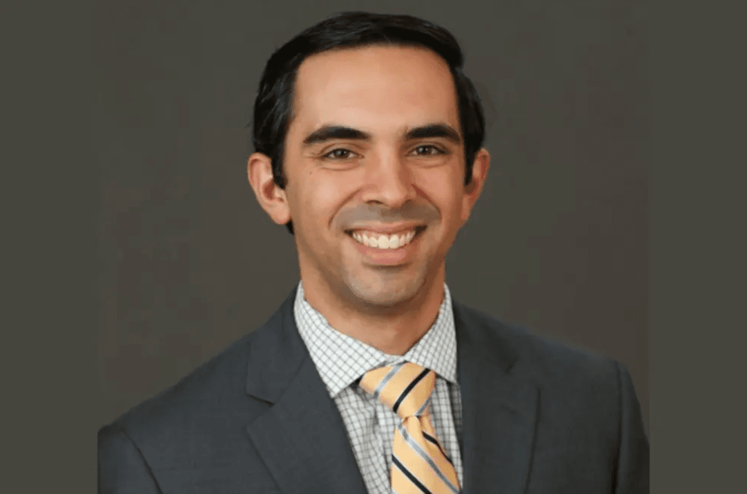 Smiling man in gray suit and yellow striped tie against a dark gray background.
