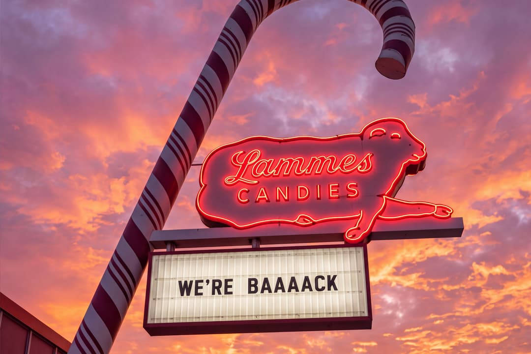 A giant red-and-white striped candy cane pole rises beside a neon Lammes Candies sign shaped like a lamb. The marquee below reads "We're baaaack" against a pink and orange sunset sky.