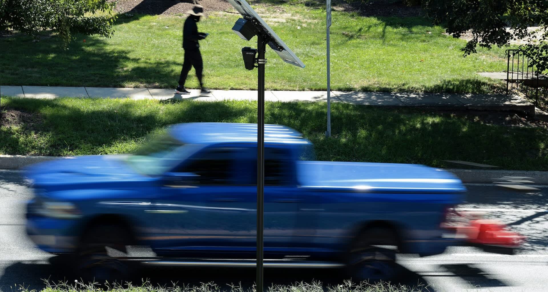 A blue pickup truck drives past a Flock Safety camera with a solar panel while a person walks on the sidewalk.