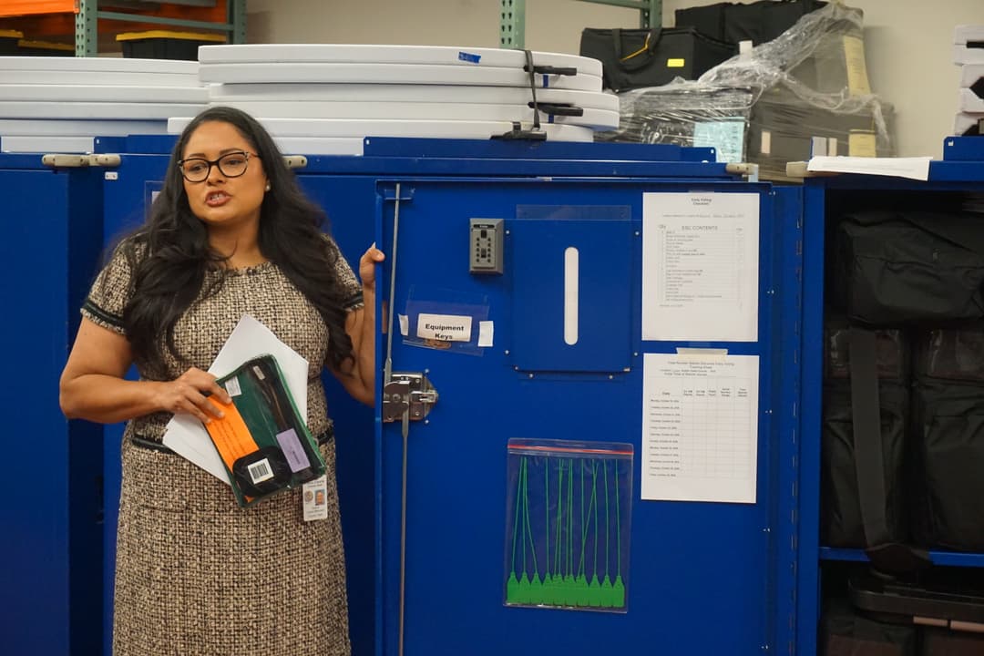 A woman speaks next to a large blue box of election equipment.