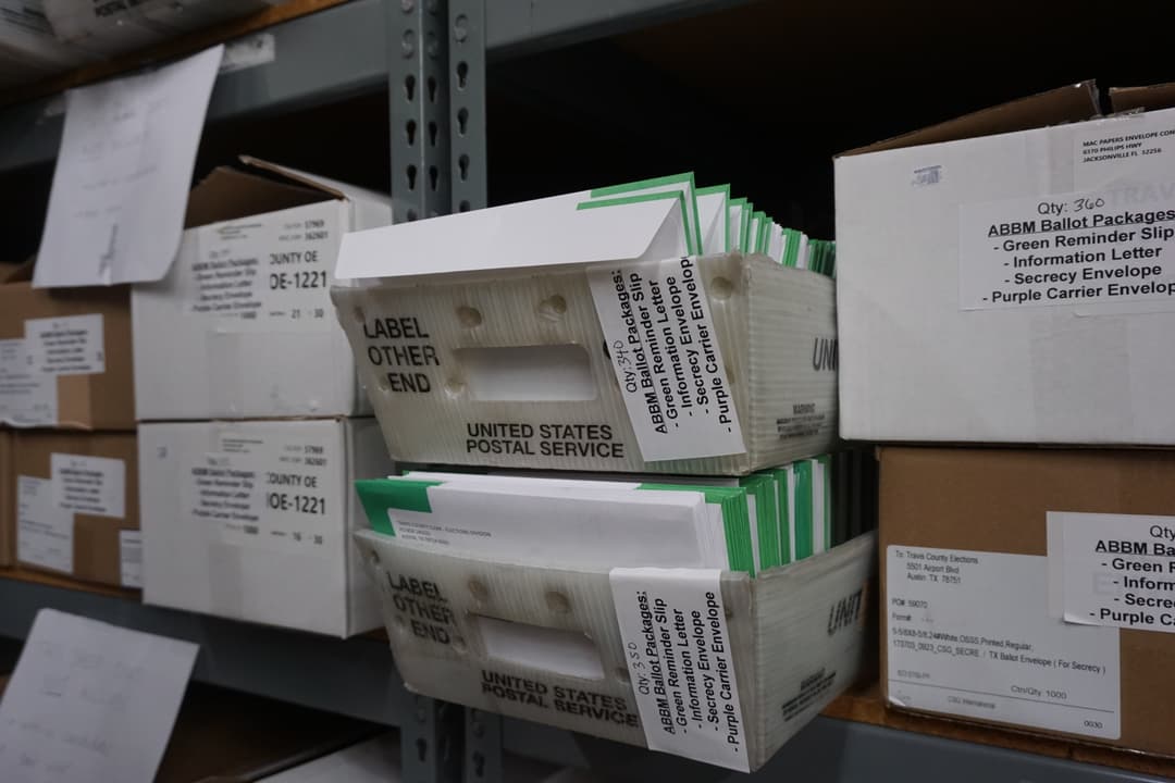 USPS trays and boxes of application for ballot by mail packages labeled for Travis County Elections on warehouse shelving.