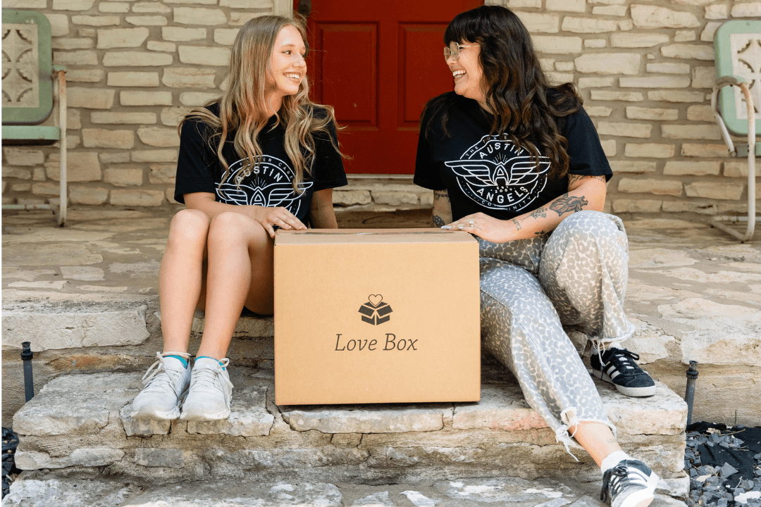 Two women in Austin Angels shirts smile at each other on stone steps beside a "Love Box" cardboard box.