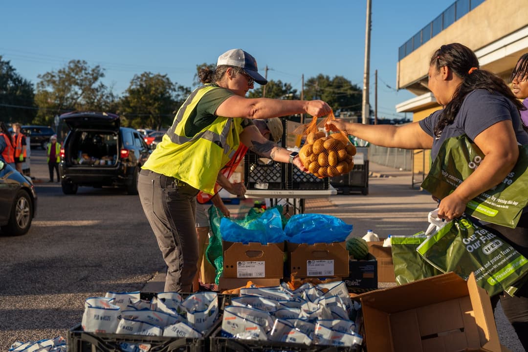 Volunteer in safety vest hands a bag of potatoes to a woman at an outdoor food distribution event.