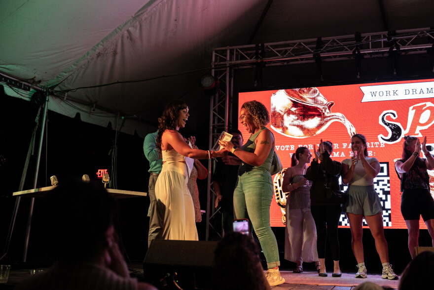 Beverlyn González hands a certificate to Amelia Johnson on stage while other performers cheer in the background.