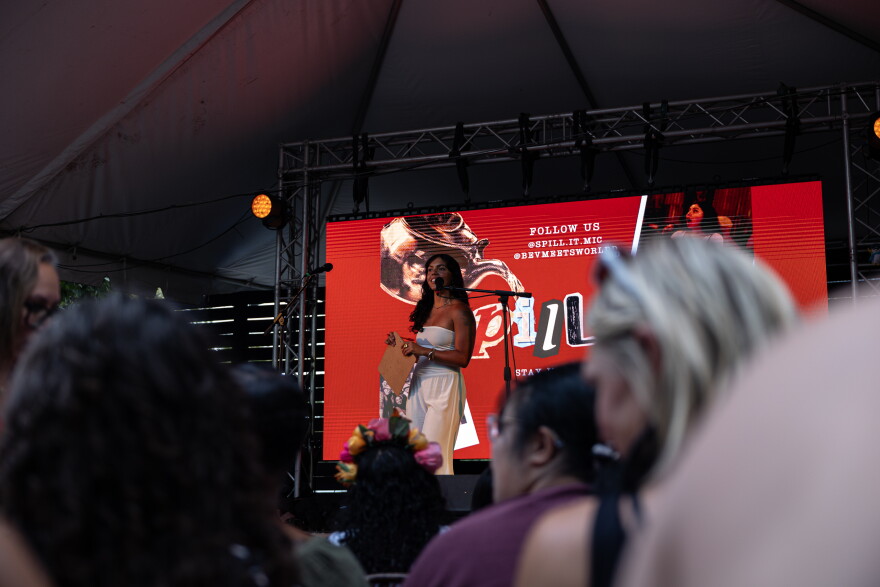 Beverlyn González speaks on stage to a crowd while holding a clipboard. The Spill It logo is on a screen in the background.