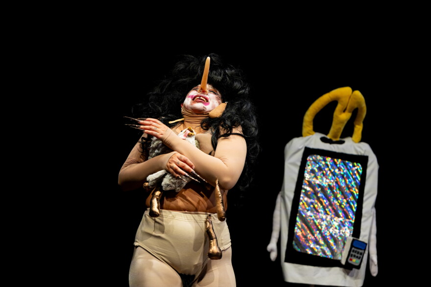 A clown-like figure with an elongated nose in a nude bodysuit holds a bird. Next to her is a fast-food ordering kiosk
