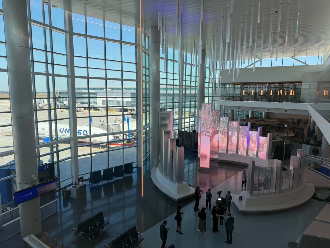 Modern airport terminal with floor-to-ceiling windows, a United jet at the gate, LED art displays, and people below.