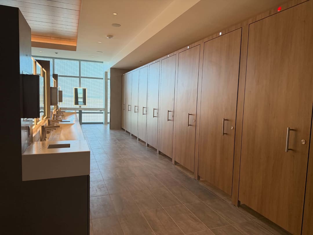 Modern restroom with floor-to-ceiling wood stall doors facing a row of sinks and lit mirrors by windows.