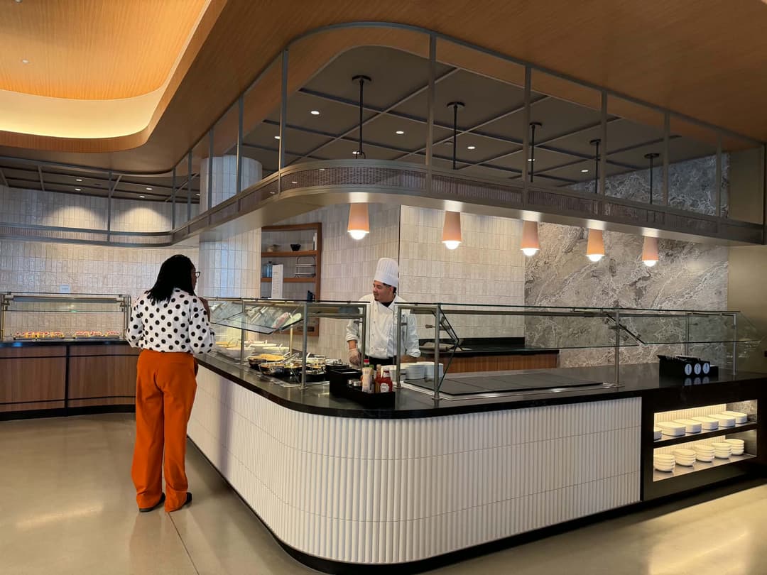 Chef in white uniform serves a customer at a modern buffet counter with food trays.
