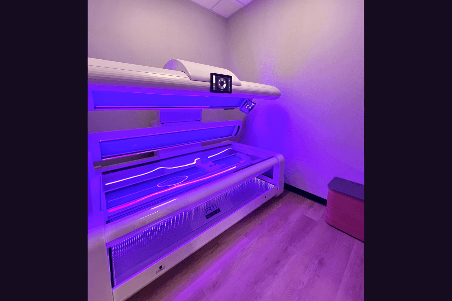 Open tanning bed glowing purple in a small room with wood floor and a cushioned stool.