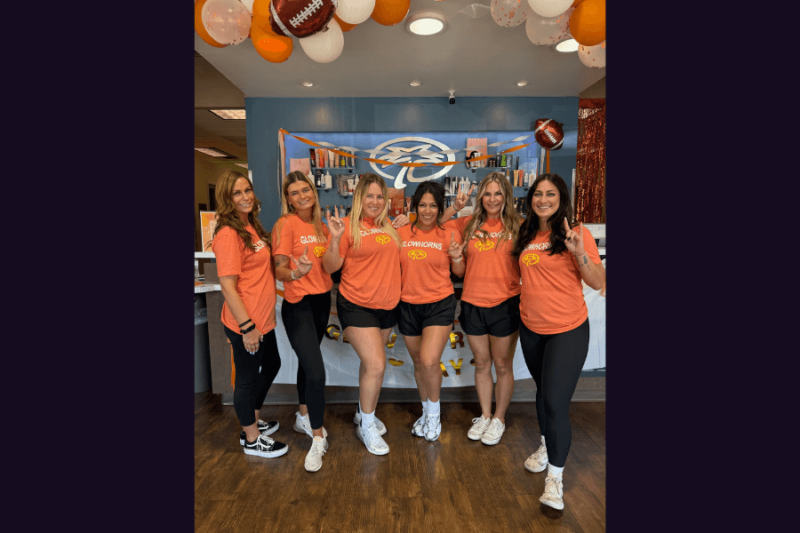 Six women in matching orange "Glowhorns" shirts pose under football-themed balloons at a salon event.