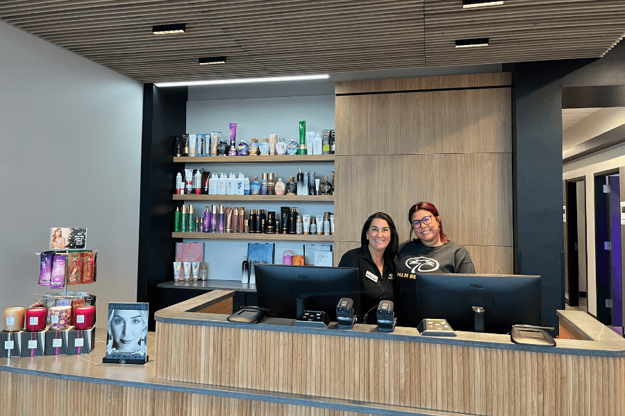 Two women smiling behind a salon reception desk with shelves of hair products and skincare items.