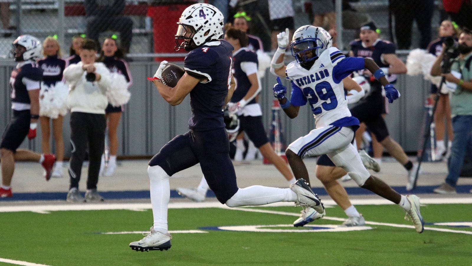 Navy-clad football player with the ball runs toward the end zone as a blue-and-white defender chases him.