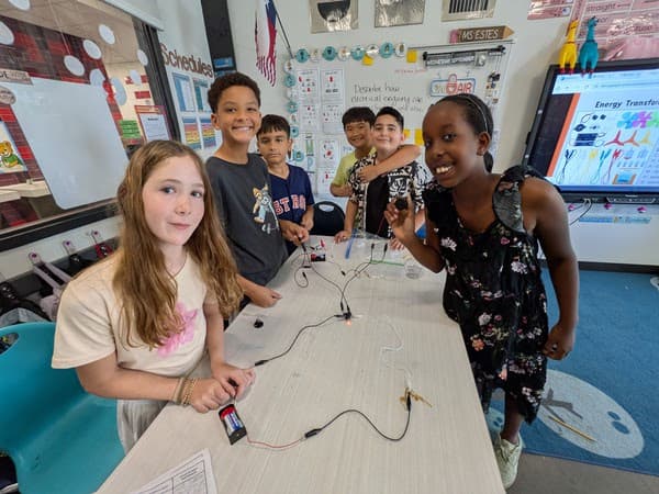 Smiling students gather around a classroom table with an electrical circuit experiment and science posters.