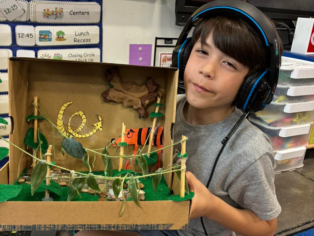 Boy wearing headphones holds a cardboard box diorama of a jungle scene with a snake, tiger, and vines in a classroom.