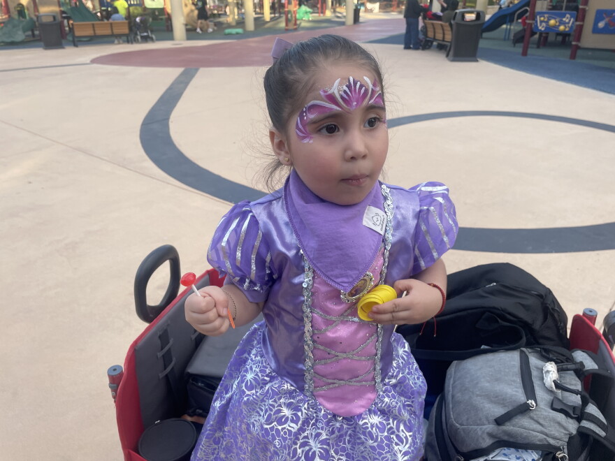 2-yr old Regina de la Rosa is dressed in a purple princess costume. Her long, straight, dark brown hair is pulled back in a tight ponytail, and vivid face paint decorates her forehead over her big brown eyes. 