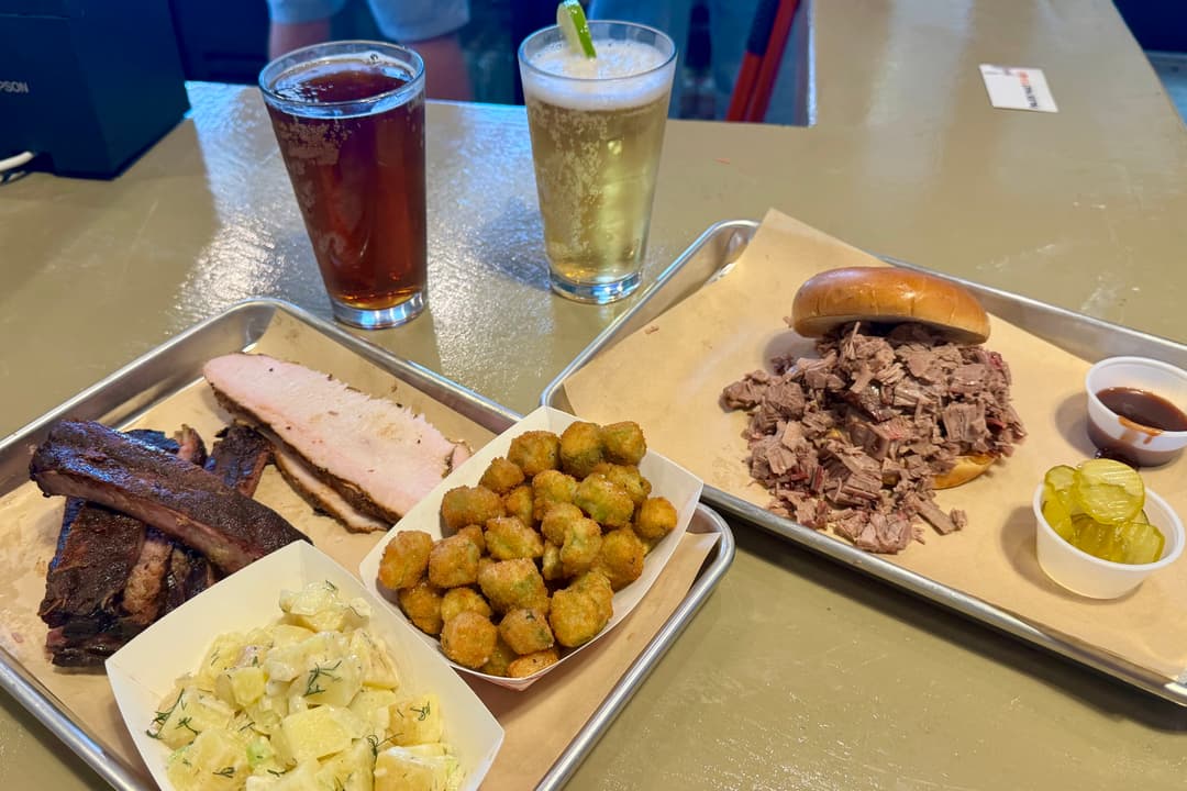 Barbecue trays with ribs, brisket sandwich, fried okra, potato salad, pickles, and two beers on a table.