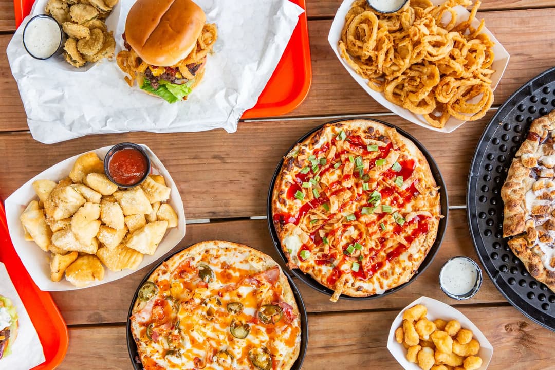 Spread of pizzas, cheeseburger, onion rings, fried pickles, breadsticks and dipping sauces on a wooden table.