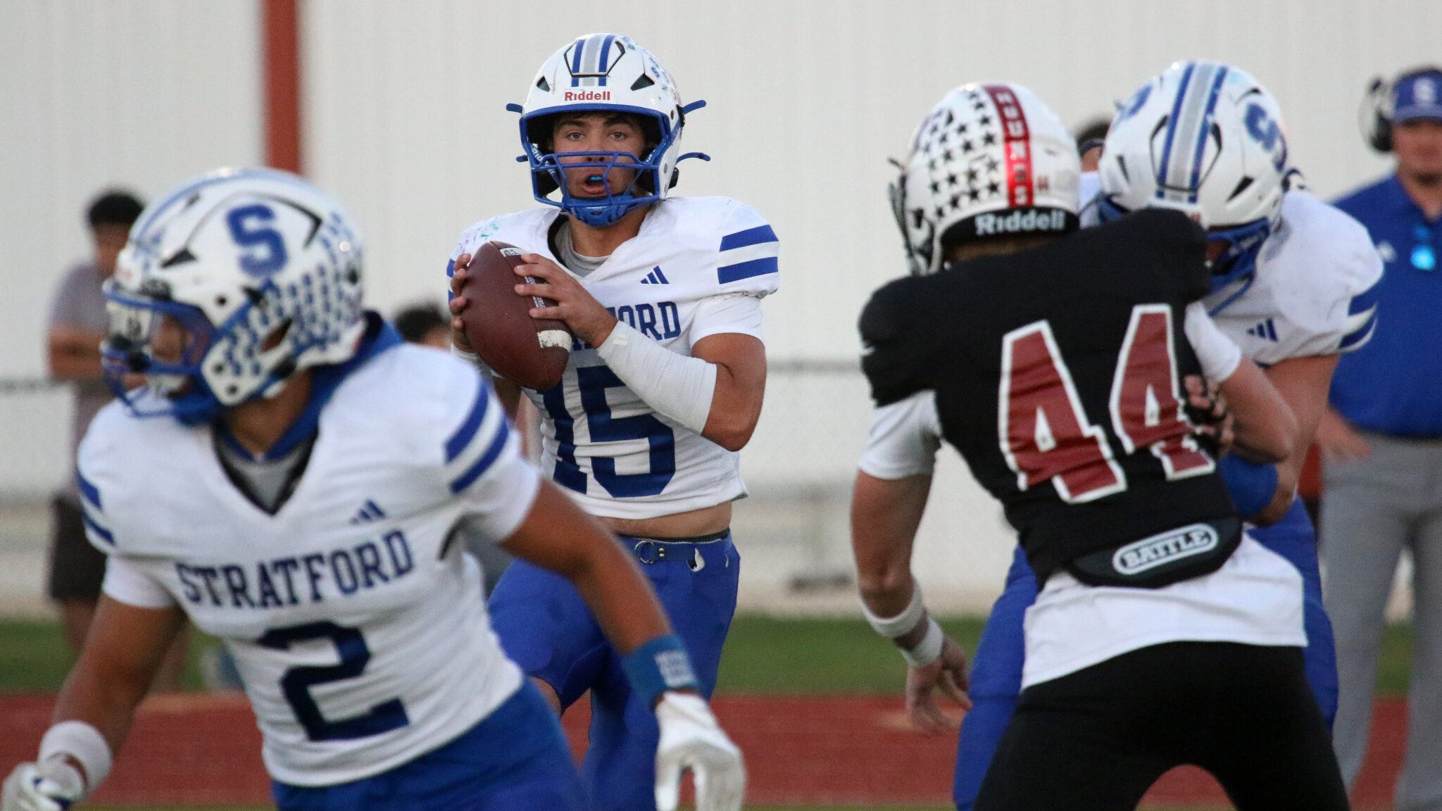 Quarterback in a Stratford white-and-blue uniform grips a football, looking to pass during a football game on a track field.