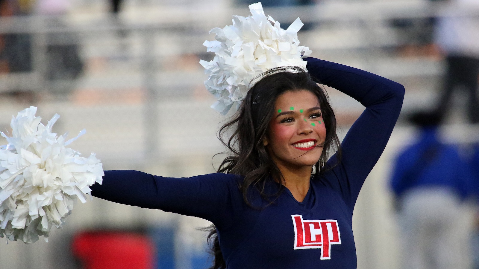 Cheerleader in a navy uniform holding white pom-poms, smiling during a performance at a stadium.