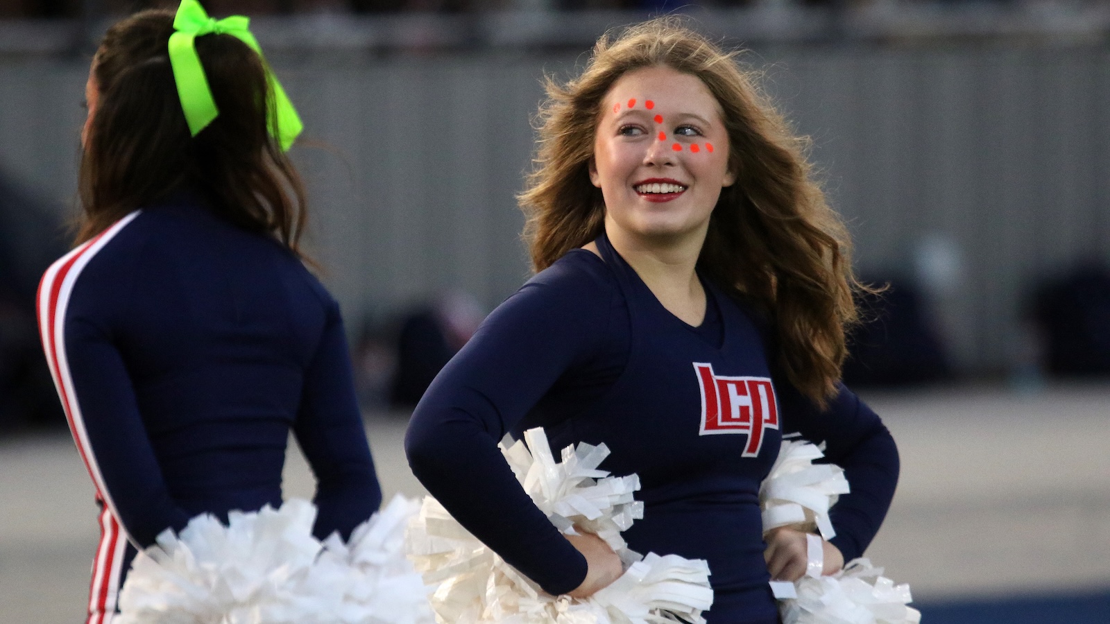 Cheerleader in navy uniform smiling, facing camera, with orange face paint dots and white pom-poms at her sides—outdoor field backdrop