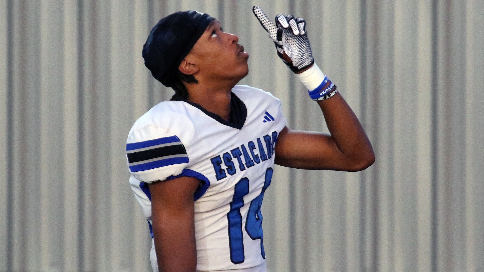 Female football player in white and blue uniform number 14, looking up with gloved hands raised near her face.
