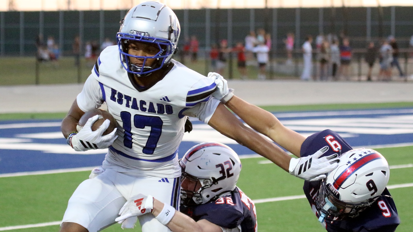 Football player in white and blue Estacado jersey (#17) runs with the ball while being tackled by two opponents on a green field with a blue end zone line behind him.