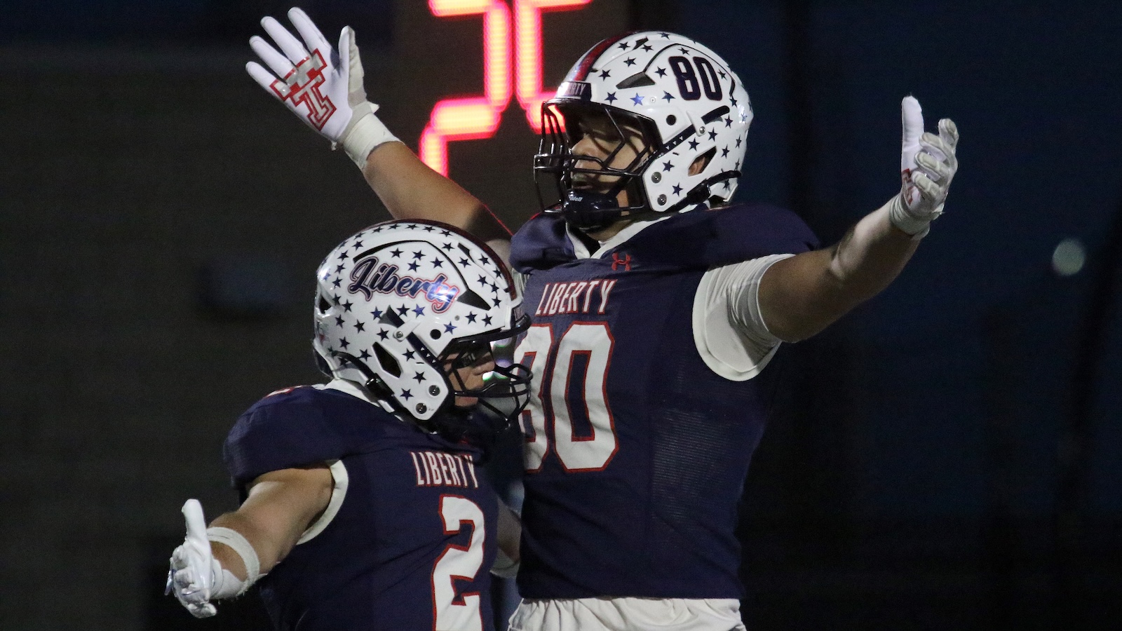 Two Liberty football players in navy uniforms celebrate with arms raised at night under stadium lights.