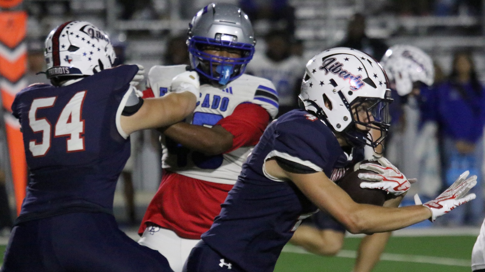 Football action at night: a navy defender wearing #54 engages a white-uniformed opponent while another navy player closes in on the ball carrier.