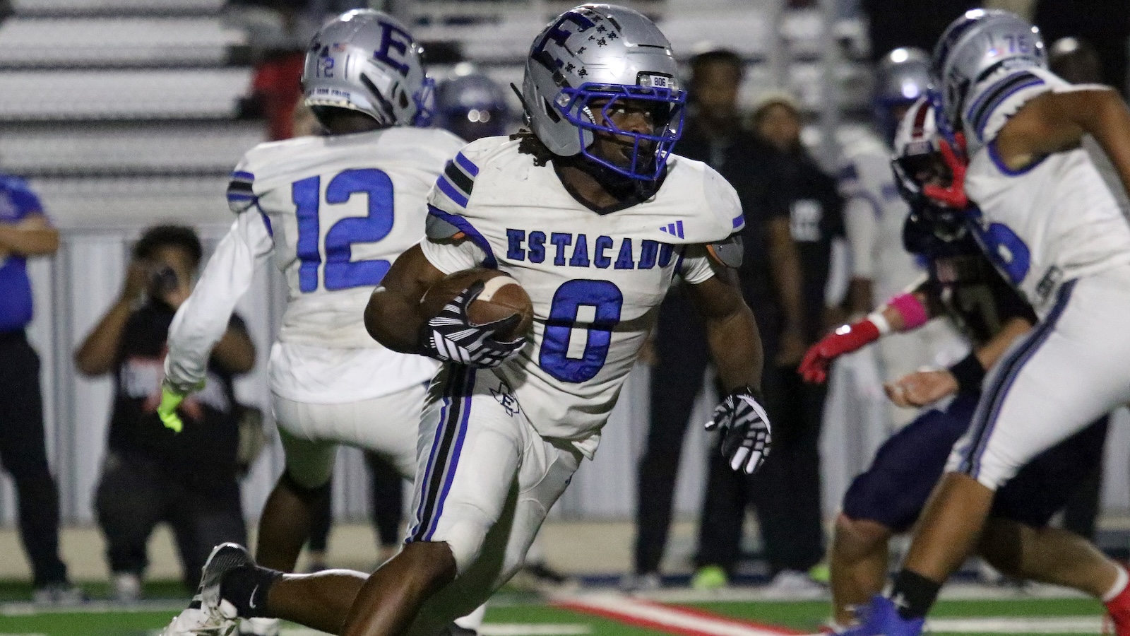 Estacado football player No. 0 runs with the ball during a game, sprinting past defenders in white and blue.