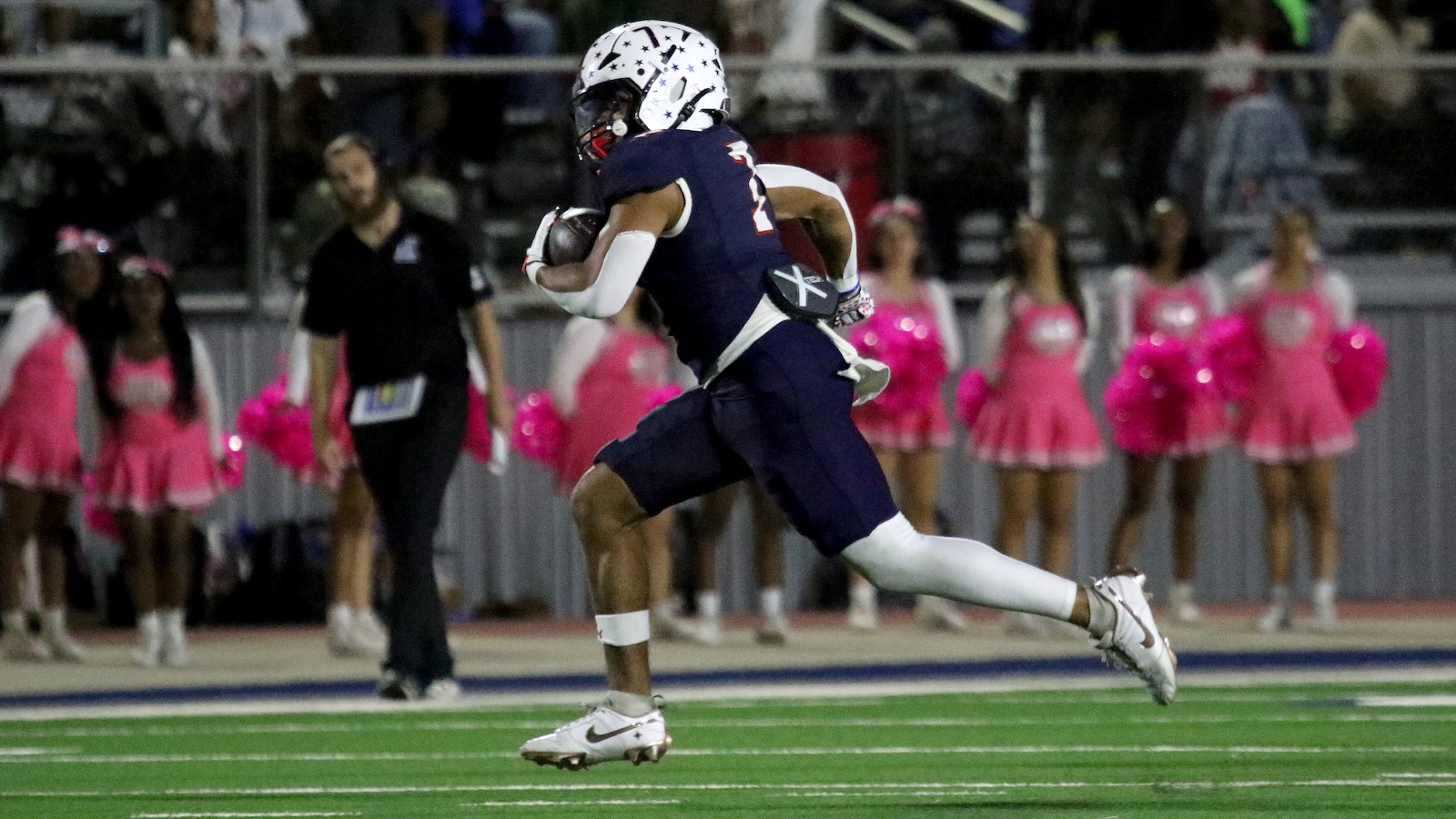 Football player in navy uniform with star-pattern helmet runs with the ball on a green field; pink-jersey cheerleaders line the sideline in the background.