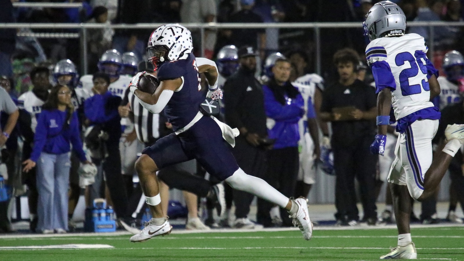 Navy-clad football player races downfield with the ball while a defender in white and blue pursues, crowd watching in the background.
