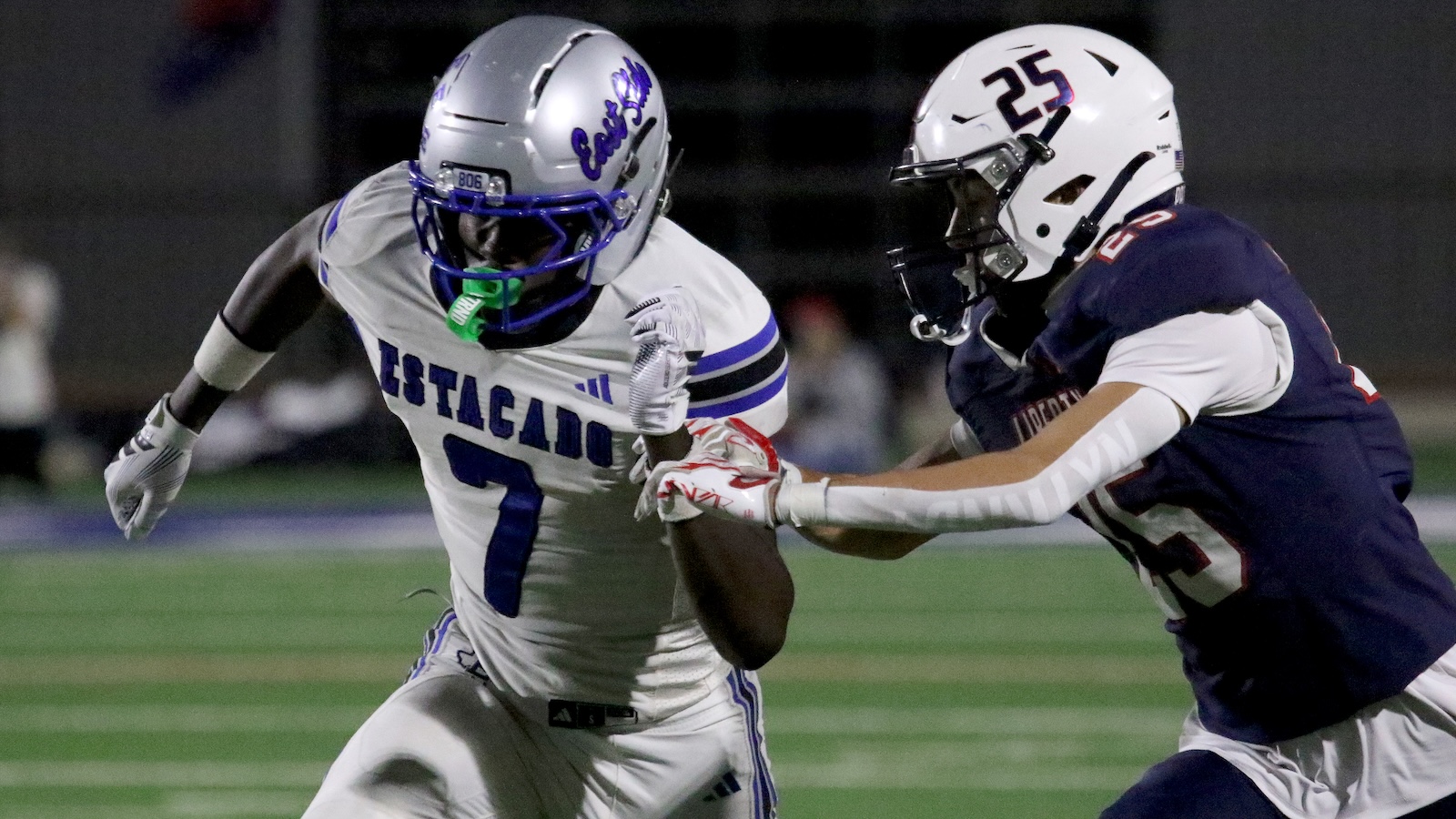 Football players in a night game: a white-jersey runner (#7, ESTACIO) is pressed by a defender in navy (#25) on a green field.