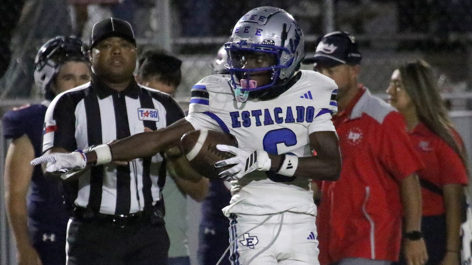 American football player in a white ESTACADO uniform with a blue helmet carries the ball as a referee signals nearby.
