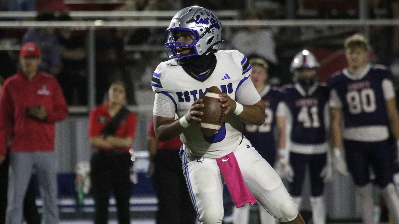 Football quarterback in white and purple uniform drops back to pass while clutching a brown football, pink flag belt trailing from his waist, under stadium lights.