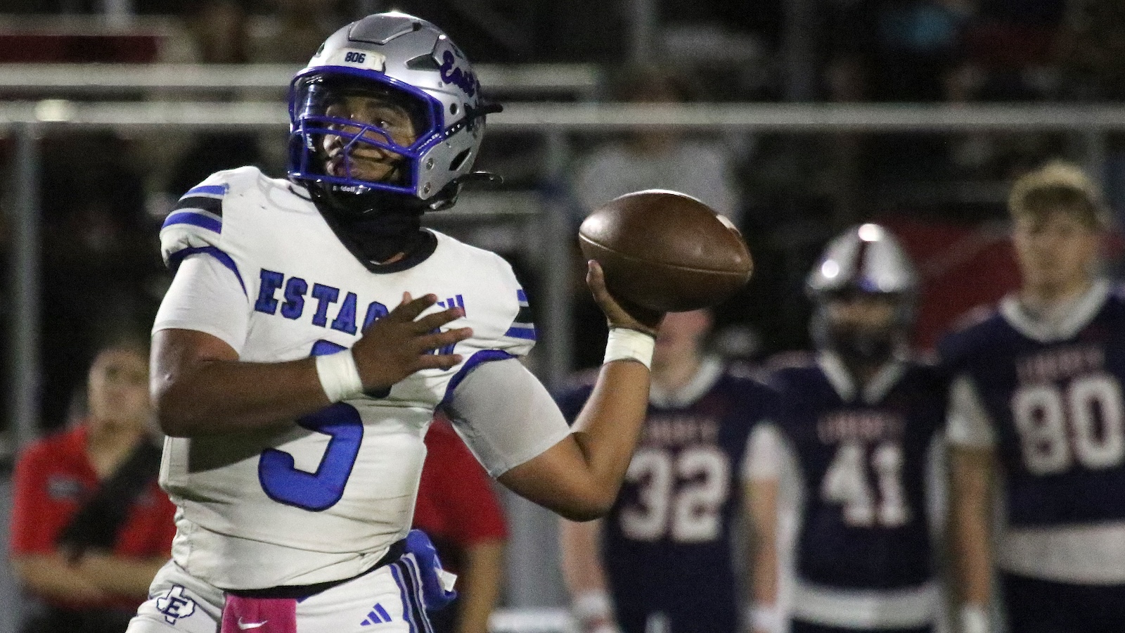 Football player in a white and blue uniform reaches to trap a spiraling football during a night game.